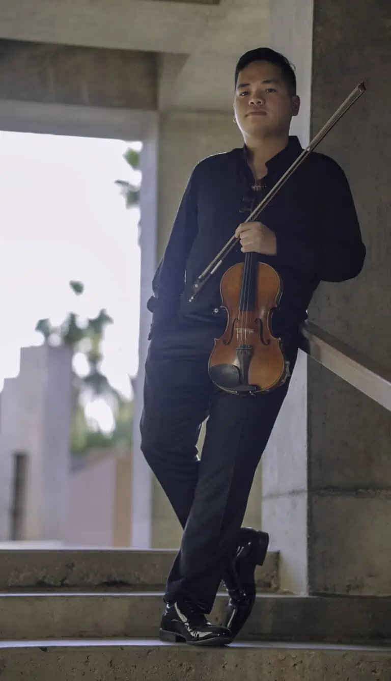 Young man in black dress shirt and slacks holding a violin and bow on a set of stairs outside.