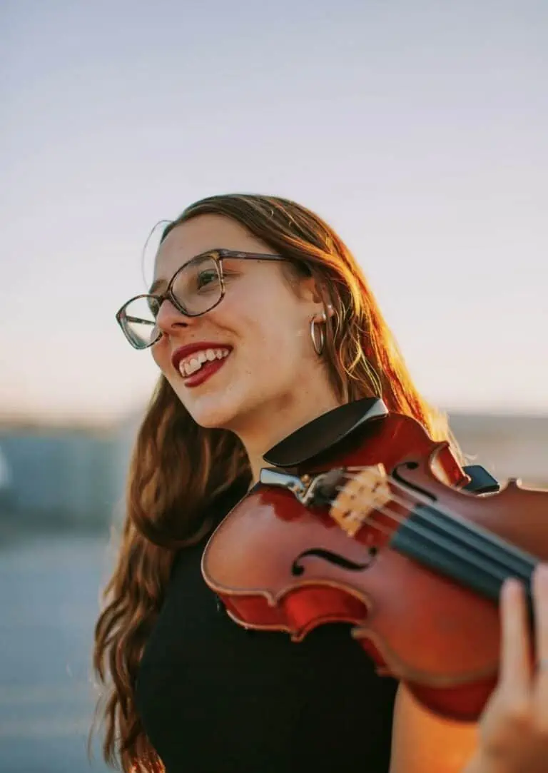 Megan Frederick headshot (young woman with long red/brown hair and glasses, smiling) holding a violin and looking away from the camera.