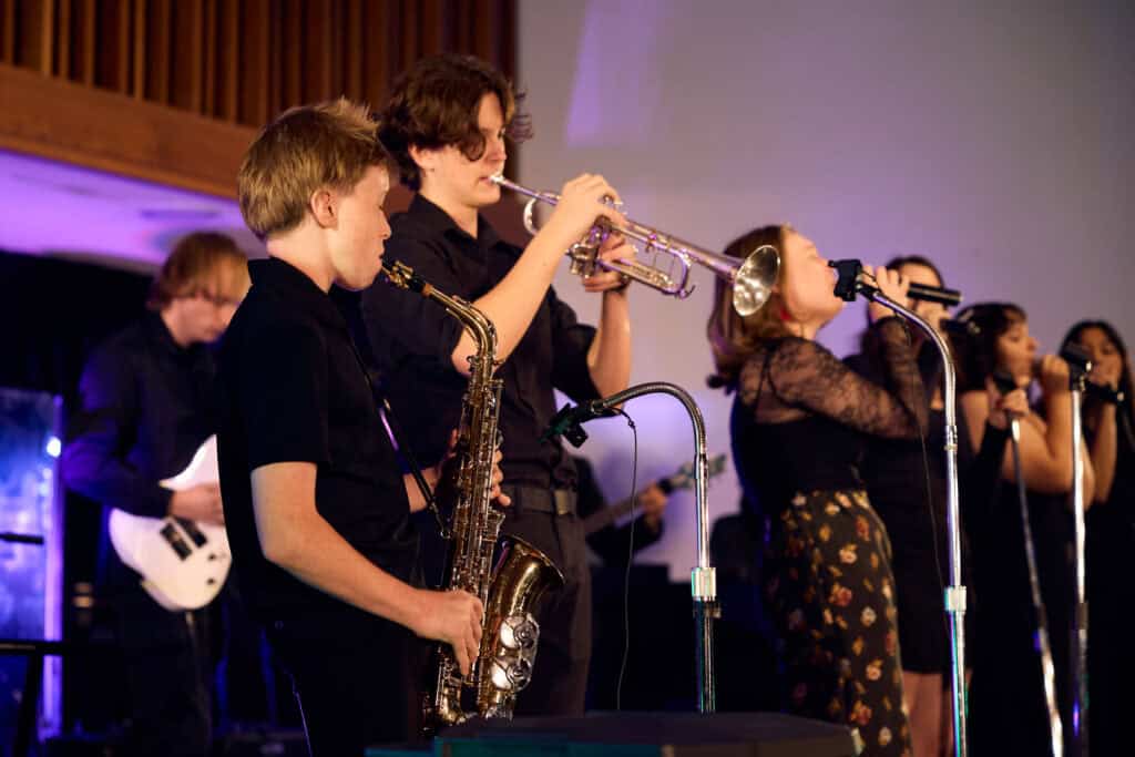 Group of students playing music on stage showing an alto saxophone, trumpet and singers.