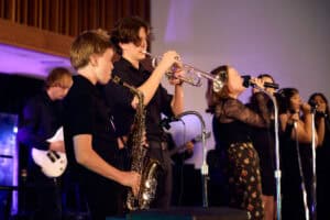 Group of students playing music on stage showing an alto saxophone, trumpet and singers.