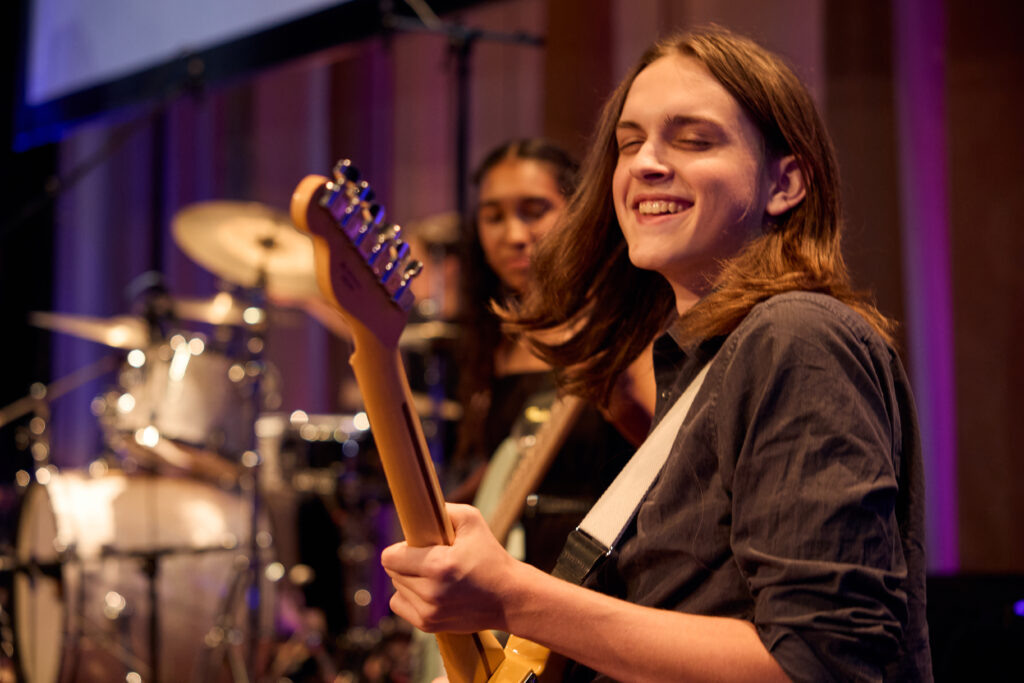 Student, Patrick Cassidy, playing guitar and smiling while bandmates look on.